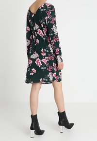 Bloemige jurk met een groene achtergrond, lange mouwen, v-hals en elastische taille; versierd met roze en witte bloemen. Gecombineerd met zwarte enkellaarsjes.