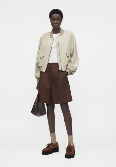 Γυναίκα με μπεζ μπουφάν bomber, λευκό πουκάμισο, καφέ σορτς μήκους μέχρι το γόνατο, μπεζ κάλτσες, καφέ παπούτσια πλατφόρμας, κρατώντας καφέ τσάντα.