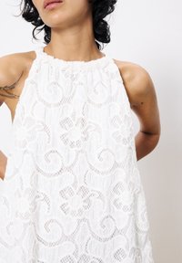 Weißes Spitzen-Haltertop mit floralen und spiralmuster, texturiertem Stoff, gerafftem Ausschnitt und offenem Rückendesign.