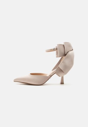 Beige pointed-toe high heel shoe with ankle strap and large padded bow detail on heel, displayed against plain light background.