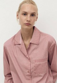 Camicia button-up rosa chiaro con colletto, maniche lunghe e taschino sul petto. Tessuto liscio con un aspetto leggermente stropicciato.