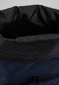 Modrý batoh s černým vnitřkem a šňůrkovým uzávěrem. Materiál má texturovaný povrch, zdobí ho vzor s logem a plastové zapínání.