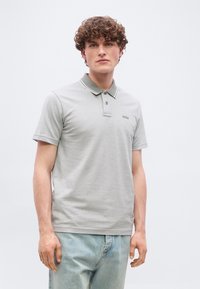Jeune homme aux cheveux bouclés portant un polo à manches courtes gris clair avec le logo "BOSS" et un jean bleu clair, debout devant un fond uni.
