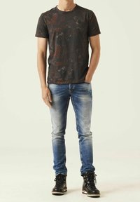Muži v tričku s černým a hnědým vzorem, vybledlých modrých džínách a černých šněrovacích botách, stojící s jednou rukou v kapse.