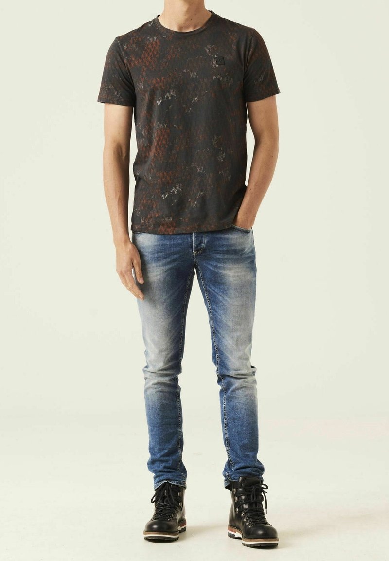 Muži v tričku s černým a hnědým vzorem, vybledlých modrých džínách a černých šněrovacích botách, stojící s jednou rukou v kapse.