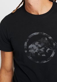 Camiseta de algodón negra con un diseño de logo circular texturizado y reflectante que presenta un motivo de elefante en tonos de gris en la parte frontal.
