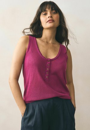 Fuchsia sleeveless top with a v-neck and five metallic button accents; made of a textured fabric. Styled with dark trousers.
