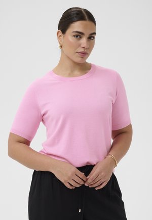 Kvinde med mørkt hår iført en lyserød kortærmet skjorte og sorte bukser, der holder frontsnørebåndene mod en lysegrå baggrund.