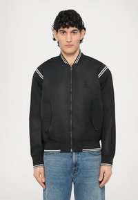 Fekete bomber dzseki fehér csíkokkal a vállakon, elöl cipzárral és két oldalzsebbel. Sima anyagból készült, bordázott mandzsettákkal és szegéllyel.