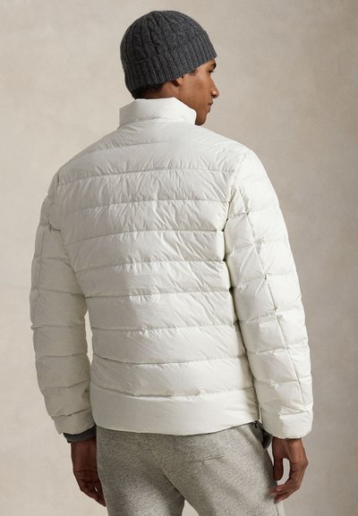 Biela prešívaná bunda s vodorovným prešívaným dizajnom, vysokým golierom a elastickými manžetami. Nosená so sivou pletenou čiapkou a sivými nohavicami, zobrazená zozadu.