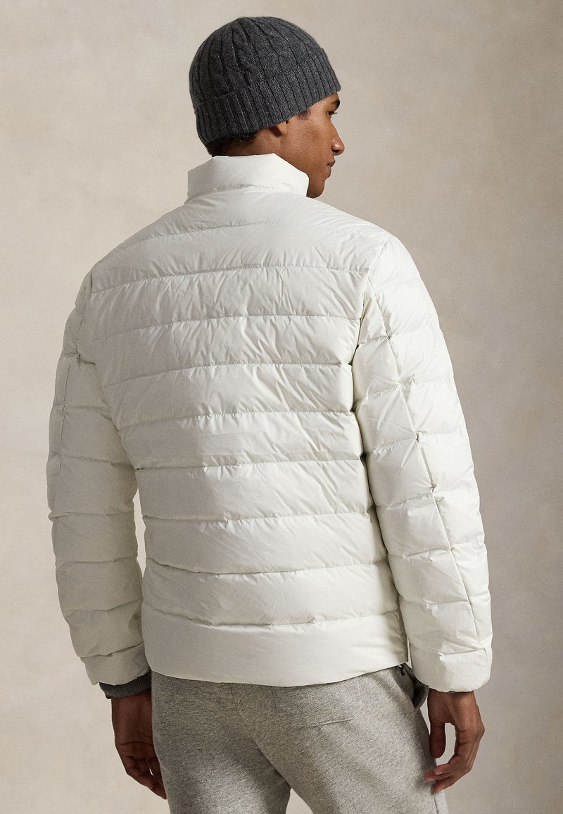 Doudoune blanche à motifs matelassés horizontaux, col montant et poignets élastiques. Portée avec un bonnet gris tricoté et un pantalon gris, vue de dos.