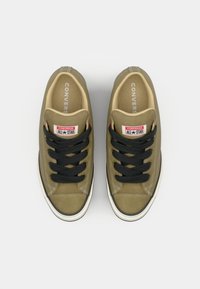 Olive green canvas sneakers with black laces, white rubber sole, and red and white "Converse All-Star" branding on the tongue.