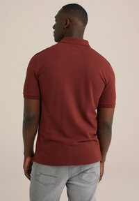 Tricou polo roșu închis cu mâneci scurte, design ajustat, material texturat și guler subtire cu dungi. Asortat cu blugi gri deschis.