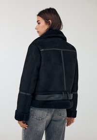 Črna umetna suede jakna z ovčjim ovratnikom, usnjenimi detajli in pasom. Ima strukturirano zasnovo in teksturirane manšete.