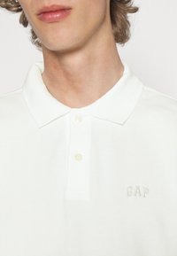 Hvid polo skjorte lavet af tekstureret stof, med en to-knap placket og subtil GAP-logo broderet på brystet i lys grøn.