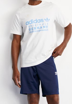 Mężczyzna ubrany w białą koszulkę Adidas Originals z niebieskim napisem "GERMANY 91074 Herzogenaurach" oraz granatowe spodenki Adidas z białym logo i paskami.