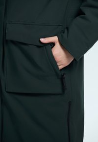 Ciemnozielony długi płaszcz z dużą przednią kieszenią, gładką teksturą i suwakową kieszenią boczną, o luźnym kroju i widocznym szwie.