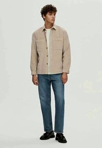 Kihara mies, joka on pukeutunut beige raidalliseen button-up-paitaan valkoisen T-paidan päällä, sinisiin farkkuihin ja mustiin kenkiin, seisoo tasaisella taustalla.