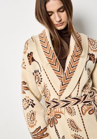 Beżowy sweter z dzianiny, zdobiony skomplikowanymi brązowymi i czarnymi wzorami paisley, z szerokim kołnierzem i pasującym paskiem z zygzakowatym wzorem.