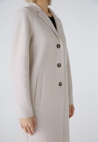 Hellbeige Wollmantel mit Reverskragen, drei Knöpfen, seitlichen Taschen und strukturiertem Stoff. Lange Ärmel mit tailliertem Schnitt.