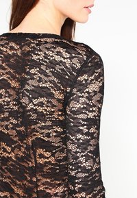 Svart spetsblus med långa ärmar, intrikata blommönster och ett genomskinligt material. Rundad halsringning som framhäver det delikata tyget.