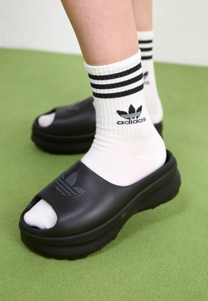 Fødder iført hvide Adidas strømper med sorte striber og sorte Adidas klipklappere på grøn gulv.