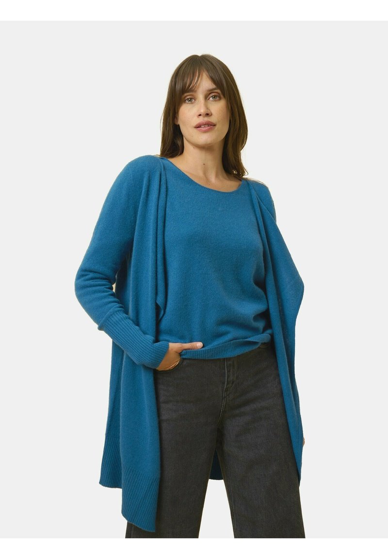 Turquoise breipullover met lange mouwen, open voorkant en geribbelde manchetten, gelaagd over een bijpassend crew-neck trui en zwarte spijkerbroek.