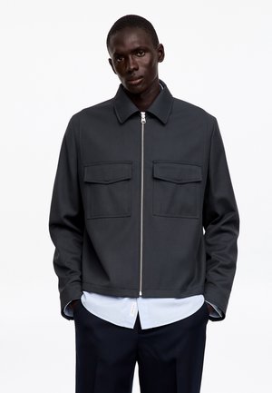 Homme portant une veste zippée gris foncé avec deux poches poitrine sur une chemise bleu clair et un pantalon foncé, debout devant un fond blanc uni.