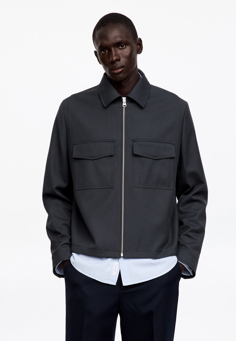 Homme portant une veste zippée gris foncé avec deux poches poitrine sur une chemise bleu clair et un pantalon foncé, debout devant un fond blanc uni.