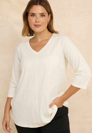 Femme aux cheveux bruns portant un haut blanc à manches trois quarts avec un col en V et un pantalon noir, une main dans la poche, devant un fond uni.