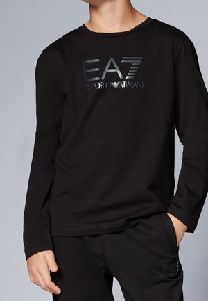 Personne portant une chemise noire à manches longues et un pantalon avec le logo brillant EA7 Emporio Armani sur la poitrine et la poche du pantalon, debout avec une main dans la poche.