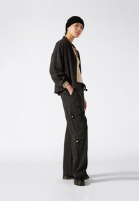 Donkergrijze button-up jack over een beige top, gecombineerd met losse donkergroene cargo broeken met zijzakken en drukknopen. Zwarte schoenen.