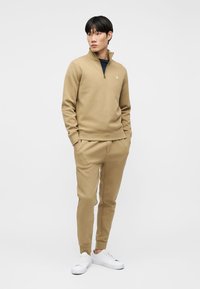Homme portant un sweat-shirt à demi-fermeture éclair beige et un pantalon de jogging assorti avec des baskets blanches, debout les mains dans les poches devant un fond blanc.