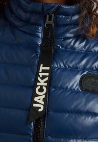 Laivastonsininen tikattu toppatakki, jossa on korkea kaulus, musta vetoketju, valkoinen "JACKIT"-merkkinen hihna ja musta merkki.