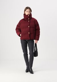 Burgunderfarbener Pufferjacke mit hohem Kragen und goldenen Knöpfen, kombiniert mit dunklen Jeans und schwarzen Stiefeletten, hält eine kleine schwarze Tasche.