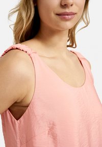 Rosa ärmelloses Top mit glatter Textur, das Rüschenträger und einen runden Ausschnitt hat. Der Stoff hat ein leicht geknautschtes Aussehen.