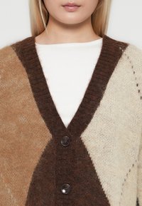 Nahaufnahme einer Person, die ein weißes Hemd unter einer braunen und beigen, weich gestrickten, geknöpften Strickjacke trägt.
