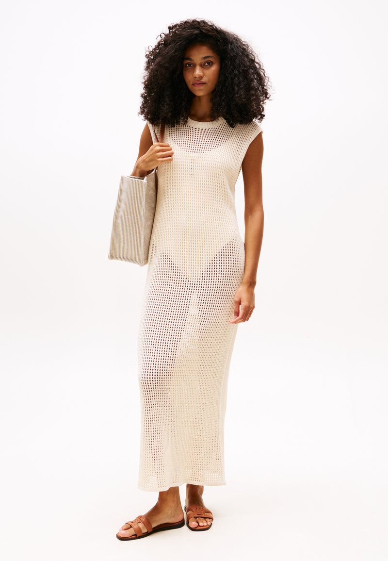 Vestido maxifino de punto beige con un diseño sin mangas, que presenta un tejido texturizado y de trama abierta. El modelo lleva un bolso tote a rayas a juego y sandalias marrones.