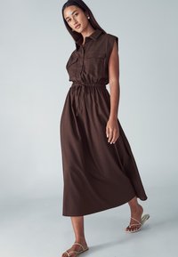 Hnedé šaty bez rukávov s golierovým výstrihom, dvoma vreckami na hrudi, elastickým pásom a plnou, voľne splývajúcou sukňou. Modelka má na sebe ploché sandále.