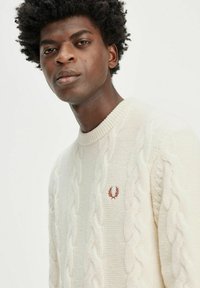 Maglione crema a trecce con collo a costine e piccolo logo ricamato sul petto. Texture morbida con motivi spessi e sollevati.