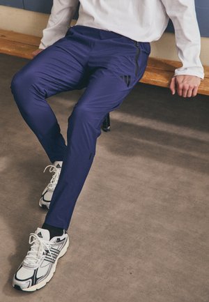 Pantalon de sport navy à coupe slim, avec des zips sur les côtés et un fini texturé. Associé à des chaussures de course blanches et noires.