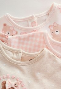 Tres camisetas para bebé en suaves tonos rosa: una con estampado de oso, una con patrón de cuadros vichy y otra con diseño de corazón y detalles bordados.