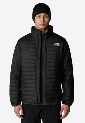 Ung mand iført en sort quiltet jakke og en matchende sort hue, stående mod en ensfarvet lys baggrund.