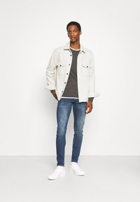 Hellgraue Knopfjacke über einem dunklen Pullover, blaue enge Jeans und weiße Sneakers. Die Jacke hat zwei Brusttaschen und schwarze Knöpfe.