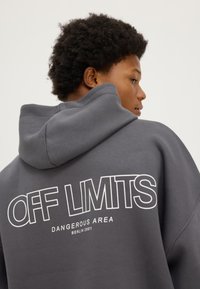 Jeune personne aux cheveux bouclés portant un sweat à capuche gris avec le texte "OFF LIMITS DANGEREUSE ZONE BERLIN 2001" dans le dos, regardant sur le côté.