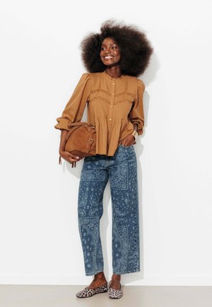 Femme avec une grande afro portant un chemisier marron, un jean à motifs bleus, des ballerines imprimé léopard et tenant un sac à main en daim marron, souriante et debout.