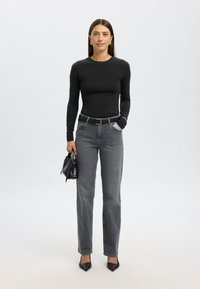 Črna majica z dolgimi rokavi, sive kavbojke z visokim pasom, črni pas, črna torbica z detajlom pentlje, črni čevlji z ostro konico, nevtralno ozadje.