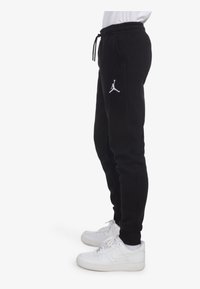Svarta träningsbyxor med elastiskt midjeband och dragsko, med en vit Jumpman-logotyp på låret. Kombineras med vita sneakers.