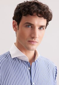 Camicia a righe blu e bianche con colletto bianco, dotata di chiusura anteriore con bottoni e texture liscia. Lo sfondo è neutro e privo di texture.