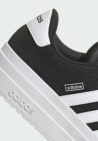 Baskets en daim noir avec des accents en cuir blanc, présentant trois bandes blanches, une semelle blanche texturée et un logo "adidas" sur le côté.